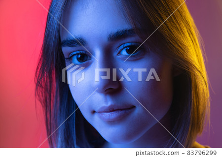 Close-up portrait of young cute girl, student looking at camera isolated on pink-purple studio backgroud in neon. Close-up portrait of young cute girl, student looking at camera isolated on pink-purple studio backgroud in neon. 83796299