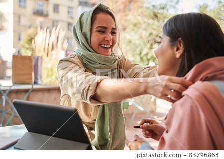 Muslim woman straightens headscarf to her girlfriend with pleasure smile Muslim woman straightens headscarf to her girlfriend with pleasure smile 83796863