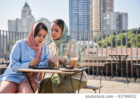 Two girl wearing abayas working attentively at the tablet while spending time at the cafe Two girl wearing abayas working attentively at the tablet while spending time at the cafe 83796932