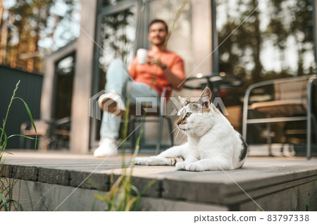 Lying cat on porch and man drinking tea behind 83797338