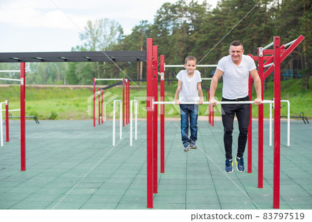 Caucasian man trains a boy on the uneven bars on the playground. Dad and son go in for outdoor sports. Caucasian man trains a boy on the uneven bars on the playground. Dad and son go in for outdoor sports. 83797519