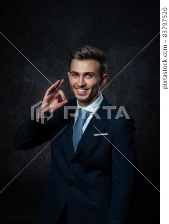 A young male steward, in a suit, smiles, makes a hand gesture everything is ok. Against a dark background. A young male steward, in a suit, smiles, makes a hand gesture everything is ok. Against a dark background. 83797520