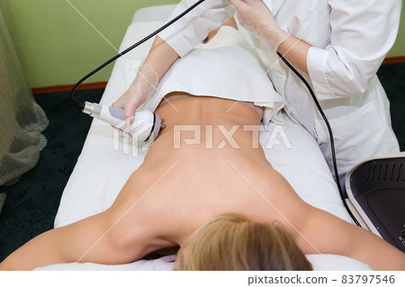 A young woman is carried out radiofrequency lifting to get rid of fat in the lateral places of the body. The cosmetologist conducts hardware cosmetology in the beauty salon. A young woman is carried out radiofrequency lifting to get rid of fat in the lateral places of the body. The cosmetologist conducts hardware cosmetology in the beauty salon. 83797546