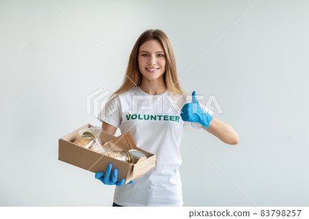 Charity organization concept. Young female volunteer holding box with food donations, showing thumb up and smiling Charity organization concept. Young female volunteer holding box with food donations, showing thumb up and smiling 83798257