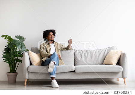 Happy Black Guy Sitting On Couch, Operating Air Conditioner With Remote Controller 83798284