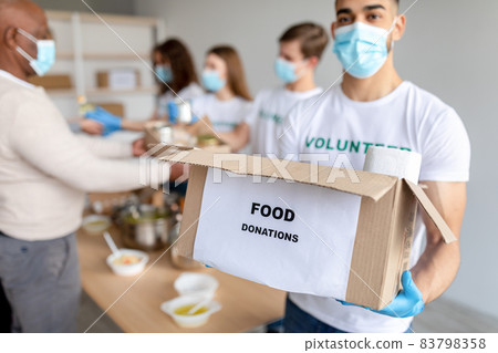 Male volunteer in medical mask holding cardboard box for donation and looking at camera, working in charity center 83798358