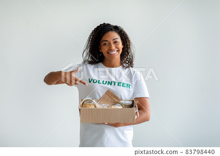 African american female volunteer holding and pointing at box with food donations, posing over light studio background African american female volunteer holding and pointing at box with food donations, posing over light studio background 83798440