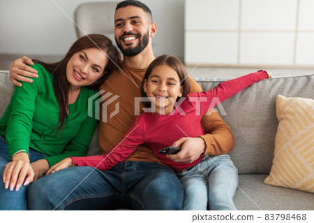 Cheerful young Arab family watching television sitting on sofa Cheerful young Arab family watching television sitting on sofa 83798468