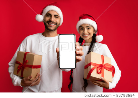 Cheerful Arab Couple Holding Christmas Presents And Showing Blank Smartphone Cheerful Arab Couple Holding Christmas Presents And Showing Blank Smartphone 83798684