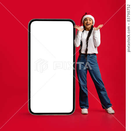 Amazing Offer. Excited Arab Woman In Santa Hat Standing Near Blank Smartphone Amazing Offer. Excited Arab Woman In Santa Hat Standing Near Blank Smartphone 83798752