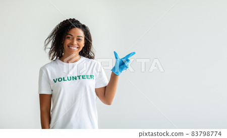 Look here. Young african american volunteer in gloves pointing aside at empty space, standing over light studio wall Look here. Young african american volunteer in gloves pointing aside at empty space, standing over light studio wall 83798774