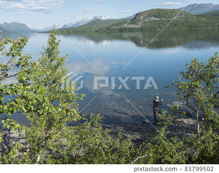 Fisherman at Lule river, in clear calm Lulealven dam lake fresh water in Saltoluokta in Sweden Lapland at sunny summer day. Green rocky mountain, birch trees and clouds. Fisherman at Lule river, in clear calm Lulealven dam lake fresh water in Saltoluokta in Sweden Lapland at sunny summer day. Green rocky mountain, birch trees and clouds. 83799502