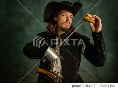 Portrait of one brutal man, medeival pirate pricking burger with sword isolated over dark background. 83801590