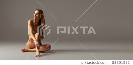 Young adorable woman in nude color underwear sitting on floor isolated over gray studio background. Natural beauty concept. 83801951