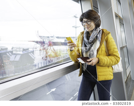 Smiling woman with passport and boarding pass is texting on her smartphone. Tourist is waiting for boarding at window at airport. 83802819