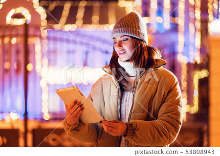 Happy young woman uses her digital tablet while standing outdoor. Christmas holidays background Happy young woman uses her digital tablet while standing outdoor. Christmas holidays background 83808943