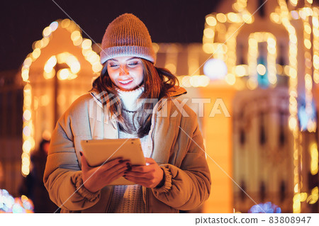 Positive girl uses her digital tablet while standing outdoor. Christmas holidays background Positive girl uses her digital tablet while standing outdoor. Christmas holidays background 83808947