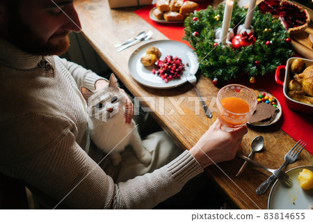 High-angle view of handsome bearded young man sitting at festive table with cat on lap and holding glass of fresh juice, during holiday family party. 83814655
