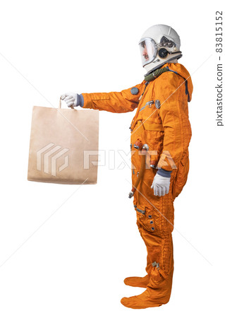Astronaut wearing orange space suit and space helmet holding in hand brown clear empty blank craft paper bag for takeaway isolated on a white background 83815152
