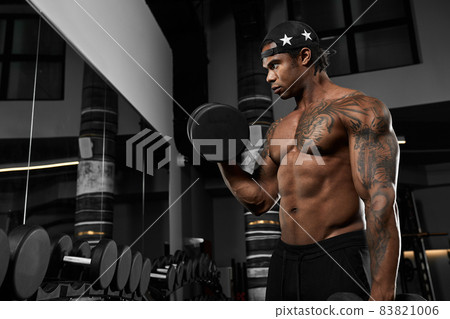 Strong and muscular dark skin man trains on modern equipment in gym. Portrait of muscular pumped up fitness trainer Strong and muscular dark skin man trains on modern equipment in gym. Portrait of muscular pumped up fitness trainer 83821006