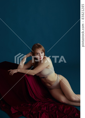 Classic portrait of young beautiful red-headed woman in nude color underwear posing isolated over blue background. 83821285