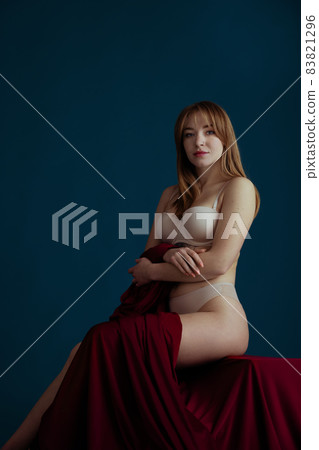Classic portrait of young beautiful red-headed woman in nude color underwear posing isolated over blue background. Classic portrait of young beautiful red-headed woman in nude color underwear posing isolated over blue background. 83821296