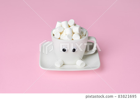 Funny and cute cup of cocoa with marshmallows. White Cup with eyes and marshmallow hat isolated on pink background. 83821695