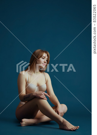 Young beautiful red-headed girl in nude color innerwear sitting on floor isolated over blue studio background. Natural beauty concept. Young beautiful red-headed girl in nude color innerwear sitting on floor isolated over blue studio background. Natural beauty concept. 83822065