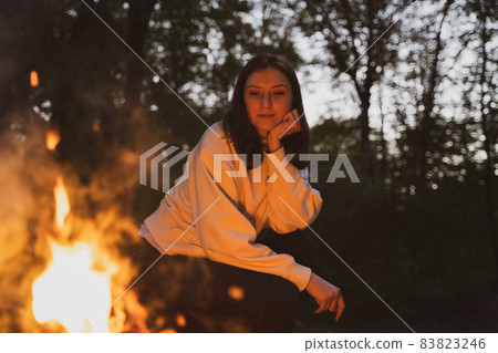A young woman sits next to a fire in a camp. Warm and cozy campfire A young woman sits next to a fire in a camp. Warm and cozy campfire 83823246