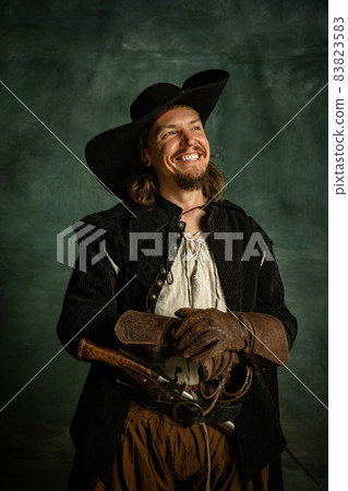 Cropped portrait of smiling man, medeival pirate in hat, gloves and cloak with pistol isolated over dark background. Cropped portrait of smiling man, medeival pirate in hat, gloves and cloak with pistol isolated over dark background. 83823583