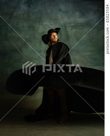 Full-length portrait of genteel brutal man, medeival pirate in hat and cloak with sword isolated over dark background. 83823584