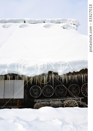 Thick snow is piled up on the roof of an old house in a snowy village, and icicles hang down. The sky is sunny. 83827018