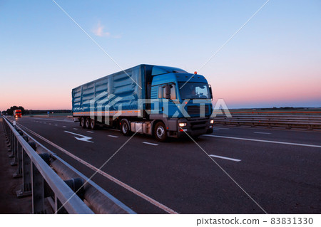 A long-distance truck driver is driving a truck with a tilt semitrailer on an evening highway. Trucking business concept, indusrty. Copy space for text 83831330