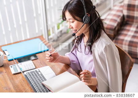 A woman wearing a headset, holding a writing instrument and facing a laptop A woman wearing a headset, holding a writing instrument and facing a laptop 83838607