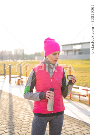 Young fit woman in warm clothes holding metal thermos drinking hot tea or coffee 83839363