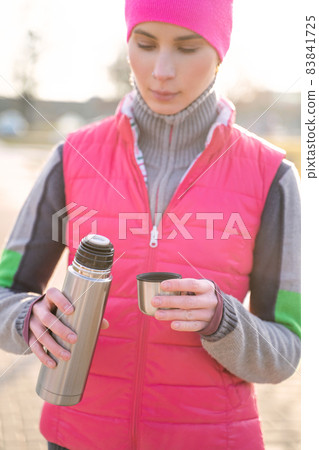 Young fit woman in warm clothes pours hot tea or coffee into mug from thermos 83841725