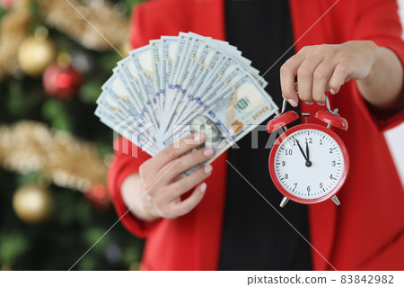 Woman holding red alarm clock and lot of american dollars near new year tree closeup 83842982