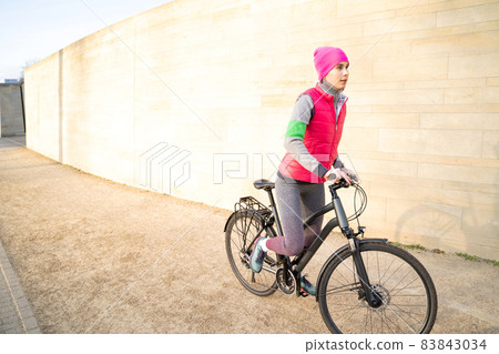 Young active woman in warm wear riding bike outdoors near wall on streets Young active woman in warm wear riding bike outdoors near wall on streets 83843034