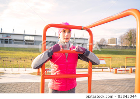 Young woman in warm fitness clothes exercises on bar on sports ground outdoors Young woman in warm fitness clothes exercises on bar on sports ground outdoors 83843044