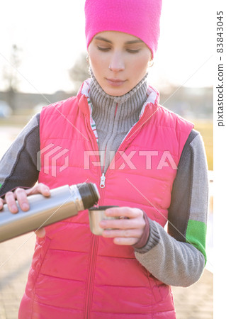 Young fit woman in warm clothes pours hot tea or coffee into mug from thermos Young fit woman in warm clothes pours hot tea or coffee into mug from thermos 83843045