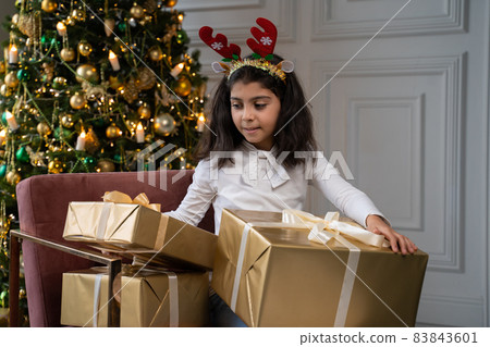 Girl is sitting on a chair next to a Christmas tree in a Christmas hat with reindeer ears and holding a gift in her hands. The child shakes the gift and wants to know what's in the box 83843601