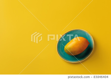 Whole ripe yellow Palmer mango on the turquoise plate over bright yellow background.  83846404