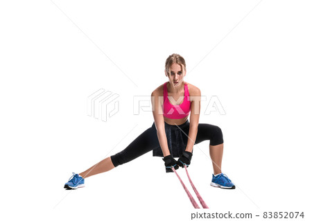 Strong woman using a resistance band in her exercise routine. Young woman performs fitness exercises on white background. Strong woman using a resistance band in her exercise routine. Young woman performs fitness exercises on white background. 83852074