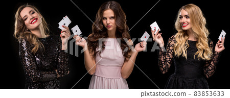 Three females in stylish dresses and jewelry. They are smiling, holding aces in their hands, posing on black background. Poker, casino. Close-up 83853633
