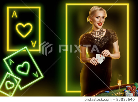 Woman in black dress showing aces, smiling, posing at table with chips, champagne. Black background with neon playing cards and frame. Poker, casino Woman in black dress showing aces, smiling, posing at table with chips, champagne. Black background with neon playing cards and frame. Poker, casino 83853640