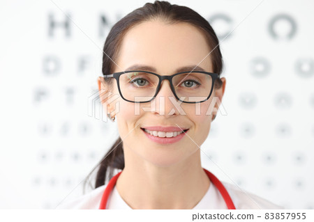 Woman ophthalmologist with glasses standing against background of table for eye test Woman ophthalmologist with glasses standing against background of table for eye test 83857955
