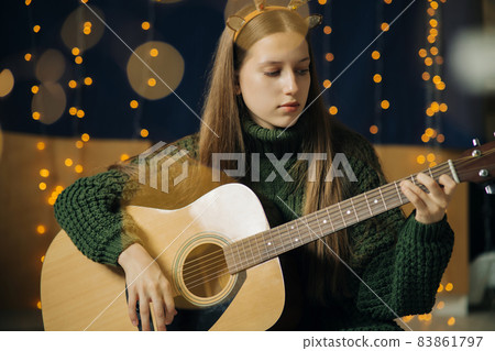 Beautiful teenage girl in Christmas antlers and sweater plays guitar 83861797