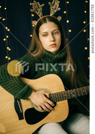 Beautiful teenage girl in Christmas antlers and sweater plays guitar 83861798