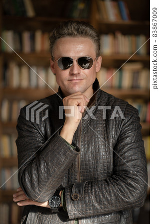 Brutal Fair-haired Guy in Modern Sunglasses Posing in his Business Office .White Man in Lather Jacket. Close-up Portrait. Book Shelves Background Brutal Fair-haired Guy in Modern Sunglasses Posing in his Business Office .White Man in Lather Jacket. Close-up Portrait. Book Shelves Background 83867009