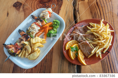 Grilled seafood skewer made of shrimps, fish and peppers with chips, carrots and broccoli, orange, on the side. On a wooden surface. Grilled seafood skewer made of shrimps, fish and peppers with chips, carrots and broccoli, orange, on the side. On a wooden surface. 83867386